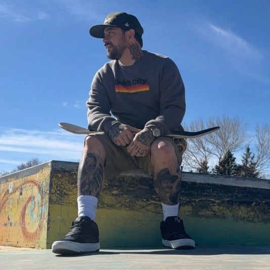 A man sitting on a concrete ledge at a skatepark, wearing a dark gray sweatshirt with a colorful 'skate city' graphic, brown shorts, black sneakers, and a black cap, holding a skateboard.